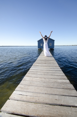 Portrait attractive mature woman standing in white long summer dress relaxed and alone with arms up on wooden boardwalk at sea, with coastline horizon and blue sky as background and copy space.の写真素材