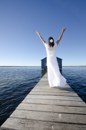 Portrait attractive mature woman standing in white long summer dress relaxed  with arms up on wooden boardwalk at sea, with coastline horizon and blue sky as background and copy space.の写真素材