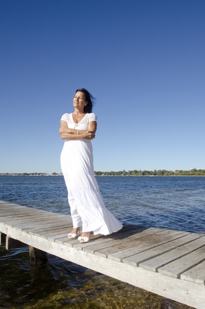 Portrait attractive mature woman standing in white long summer dress and high heel shoes relaxed  and confident on wooden boardwalk at sea, with coastline horizon and blue sky as background and copy space.の写真素材