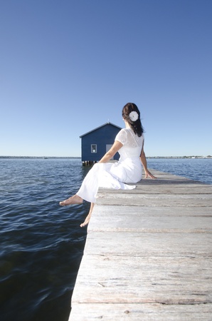 Portrait attractive mature woman sitting in white long summer dress relaxed and lonely on wooden boardwalk at seaの写真素材