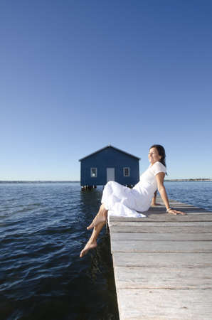 Portrait attractive mature woman sitting in white long summer dress relaxed  with closed eyes and bare feet on wooden boardwalk at seaの写真素材