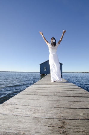 Portrait attractive mature woman standing in white long summer dress relaxed  with arms up on wooden boardwalk at sea, with coastline horizon and blue sky as background and copy space.の写真素材