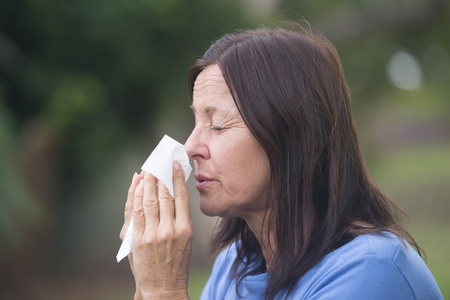 Portrait beautiful mature woman suffering from cold or flu infection, sneezing into tissue, painful seasonal hayfever, with blurred outdoor background and copy space.の写真素材