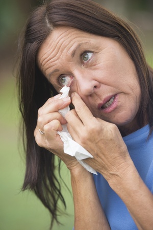 Portrait attractive sad mature woman cleaning eye with tissue, in tears or crying, with blurred outdoor background.の写真素材