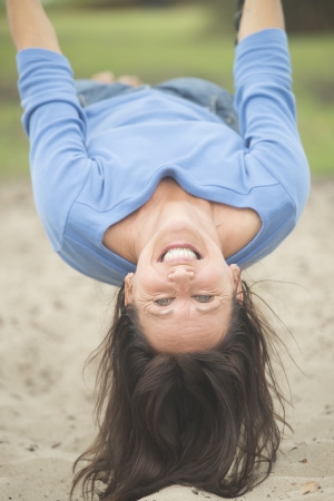 Portrait attractive mature woman hanging bare feet upside down on swing on playground, enjoying cheerful active retirement, happy smiling, with blurred outdoor background.の写真素材