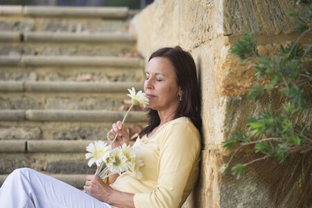Portrait attractive mature woman with happy relaxed facial expression, in love and romance with closed eyes and flower sitting at limestone wall with blurred outdoor background.の写真素材