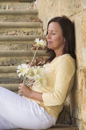 Portrait beautiful mature woman with happy relaxed facial expression, in love and romance with closed eyes and flower sitting at limestone wall with blurred outdoor background.の写真素材