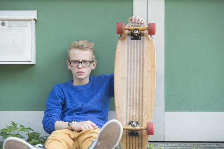 Portrait of blond teenage boy outdoor with serious face, wearing glasses and holding his skateboard.の写真素材
