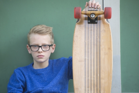 Portrait of blond teenage boy with serious face, wearing glasses and holding his skateboard.