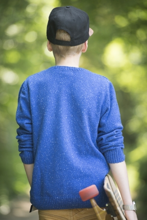 Blond teenage boy walking relaxed with skateboard under arm along alley with trees and bushes as blurred background.の写真素材