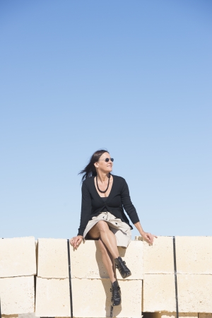 Portrait sexy mature woman with, wearing skirt, fishnet stockings and boots, is sitting confident, relaxed on wall, isolated with clear blue sky as background and copy space.の写真素材