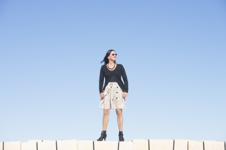 Portrait happy sexy mature woman with, wearing skirt, fishnet stockings and boots, is standing confident, relaxed and friendly on wall, isolated with clear blue sky as background and copy space.の写真素材