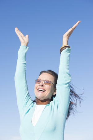 Portait beautiful looking mature woman at sunny day outdoor, with sunglasses and hands up in air, with happy joyful smile and confident gesture, isolated with blue sky as background and copy space.の写真素材