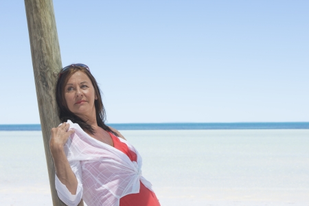 Portrait attractive mature woman on holiday leaning relaxed  at wooden pole, with tropical ocean and blue sky as blurred background and copy space.の写真素材