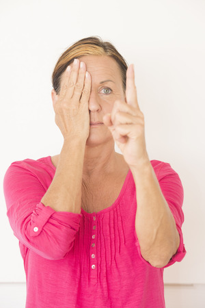 Portrait attractive mature woman in pink shirt, covering one eye with hand and focusing on finger, testing eyesight, serious face, isolated on white.の写真素材