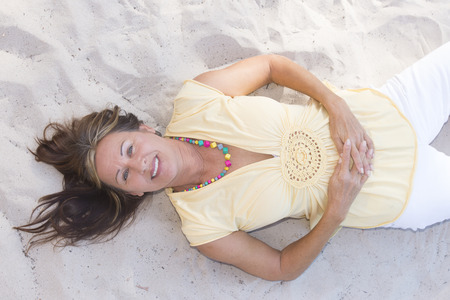 Portrait relaxed attractive mature woman lying in the sand of beach, looking up, laid back with folded hands on belly, happy smiling.の写真素材