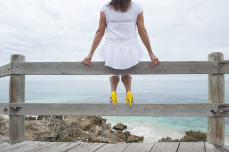 Portrait attractive mature woman sitting alone and relaxed on wooden handrail overlooking ocean, wearing white blouse and yellow high heel shoesの写真素材