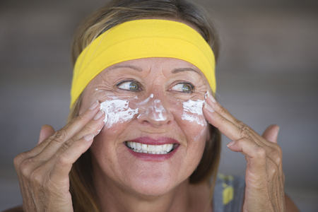 Portrait sporty fit attractive mature woman with protective sunscreen skin care creme and moisturiser lotion on joyful smiling face, yellow headband, blurred background.の写真素材