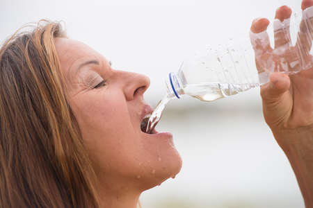 Portrait attractive mature woman enjoying a drink of mineral water as refreshment after sport activity outdoor, spills and drops on faceの写真素材