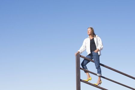 Portrait beautiful mature woman standing confident in high heel shoes on top of metal fence, laid back and relaxed, friendly smiling, with blue sky as background and copy space.の写真素材