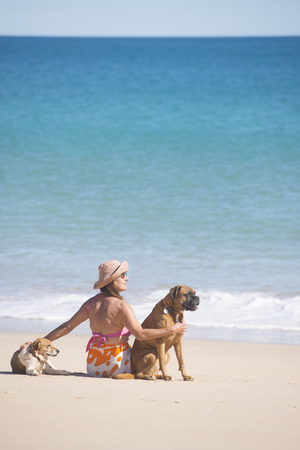 Portrait beautiful mature woman sitting happy and relaxed with dogs at beach, with ocean and blue sky as background and copy space.の写真素材