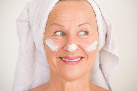 Portrait attractive senior woman with towel and protective skin care creme and moisturiser lotion on happy joyful face, bright background.の写真素材