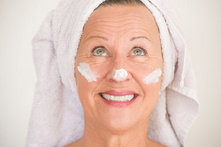 Portrait friendly attractive mature woman with towel and protective skin care creme and moisturiser lotion on happy smiling face, bright background.の写真素材