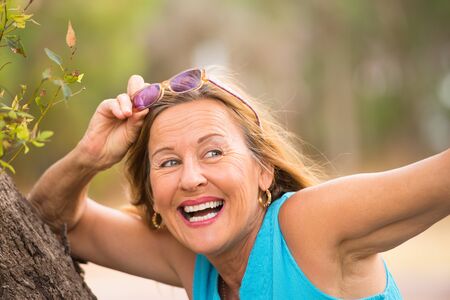 Portrait attractive mature woman with happy friendly relaxed smile and sunglasses in park, blurred outdoor background, copy space.の写真素材