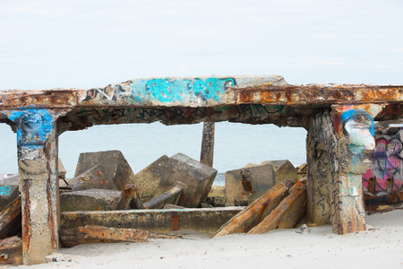 Remains of partly collapsed bridge on beach, with Scattered concrete and rusty steel debris and rubble in water and sand, graffiti, blurred background and copy spaceの写真素材