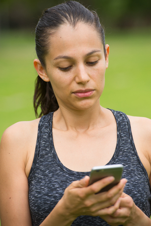 Portrait beautiful active young hispanic woman using smart phone, serious face, networking, texting, chatting in park outdoor after exercising, blurred background.の写真素材
