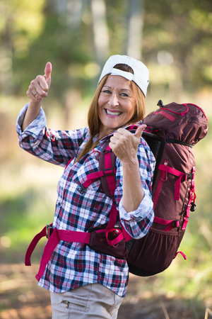 Portrait active attractive mature woman hiking happy and relaxed with backpack in the woods, thumbs up, blurred background.の写真素材