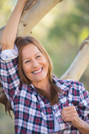 Portrait attractive mature woman in rural country, wearing plaid shirt, posing relaxed with happy smile against Australian outback bush background.の写真素材