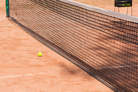 Tennis court of brown color, black tennis net and a yellow tennis ball by the net. A box of tennis balls in the background.の写真素材