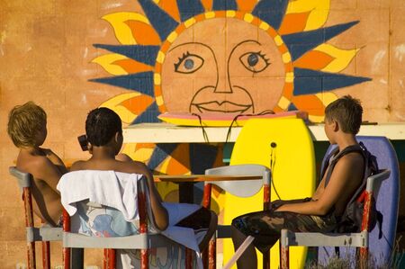 Three suntanned boogie boarder boys relaxing in front of a sunshine mural at the beach on a late summer afternoonの写真素材