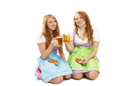 two bavarian girls with pretzels and beer kneeling on floor on white backgroundの写真素材