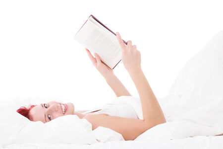 happy redhead woman in bed reading a book on white backgroundの写真素材