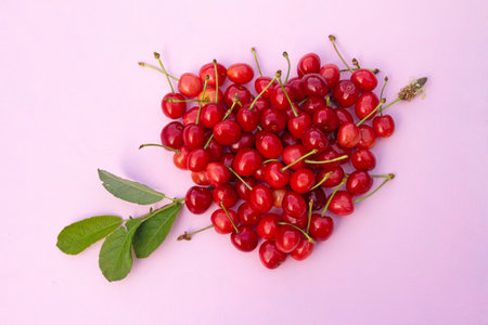 Red sweet cherries in heart shaped with arrow of green branch layout on light pink background. High...