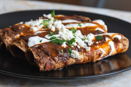 Vegetarian Mexican food, enfrijoladas. A type of enchilada dish of fresh cheese wrapped in corn tortilla and bathed in a spicy black bean sauce, topped with cream, cheese, spicy sauce and cilantroの写真素材