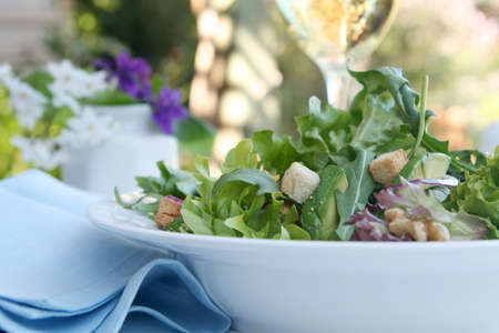 Green salad in a white bowl, outdoor setting.  With avocado, walnuts, mango, croutons and a glass of white wine.の写真素材