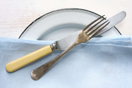 Rustic place setting, with old bone-handled knife, tarnished silver fork, enamelled plate, and blue napkin, on weathered white timber table.の写真素材