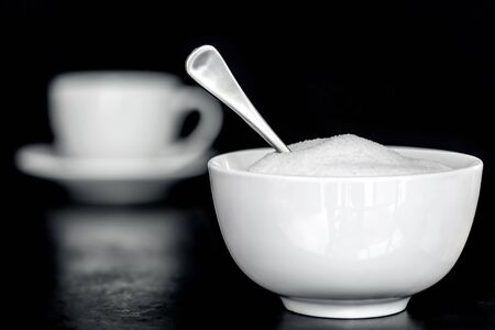 Bowl of sugar with spoon.  Coffee cup behind, over black.  Selective focus.の写真素材