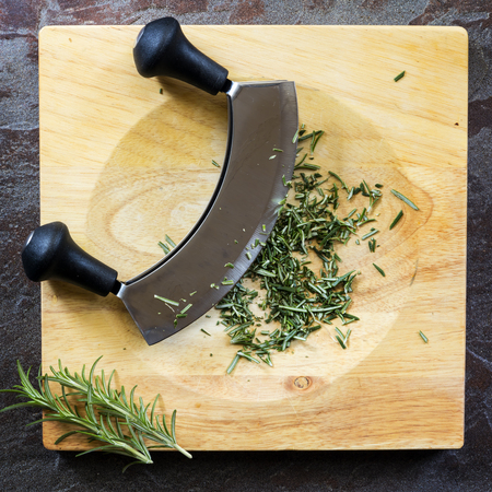 Chopping fresh rosemary with mezzaluna curved knife on board, top view on slate.