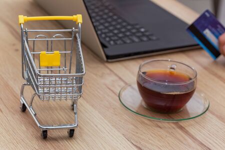 Internet shopping. Thumbnail of a shopping trolley with laptop, bank card and glass of tea. Concept. Convenient shopping without leaving your homeの写真素材