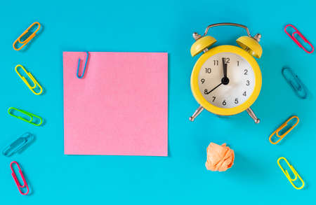 Blue background, pink card with an office staple and space for an inscription. Next to it, an alarm clock is set for an hour, five to midnight and a crumpled ball of paper. Concept for business and office purposesの写真素材