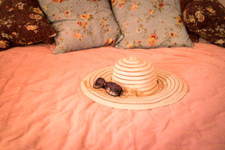 Elegant women's hat and sunglasses lying on a stylish bed. Warm light. The concept of a summer morning, vacation, pleasant relaxationの写真素材