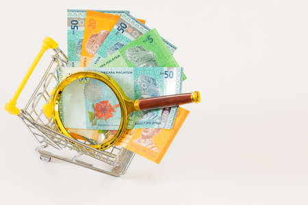 A miniature shopping cart with Malaysian currency and a magnifying glass. Economic and business concept, rising inflation and the country's economic upset. White background, Space for an inscriptionの写真素材