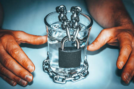 Hands reaching out of thirst for a glass of clear water closed with a padlock and a chain. The problem of climate change. Diminishing freshwater resources.の写真素材