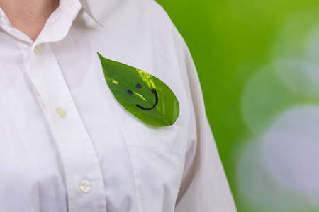 A businesswoman holds a green leaf in a shirt pocket with a painted smile, natural background with blurred greenery in the park, creative concept, development in harmony with nature, work-life balanceの写真素材