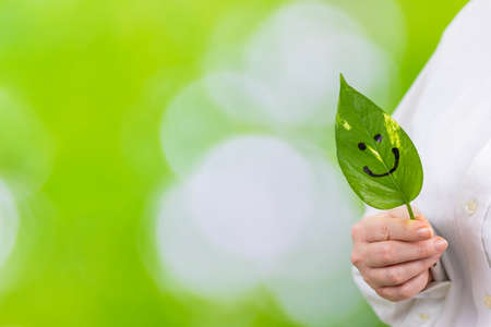 A woman holds a green leaf with a painted smile in her hands, a natural background with blurred greenery in the park, a creative idea, development in harmony with nature, work-life balance, copy spaceの写真素材