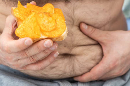 Overweight problems, Unhealthy fattening food, One hand holds a fat belly, the other holds a sandwich with chips, Concept, Obesity, Excessive consumption of saturated fatの写真素材